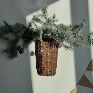 May include: A brown wicker basket hanging on a white wall with a sprig of evergreen branches with small silver ornaments hanging from the branches.