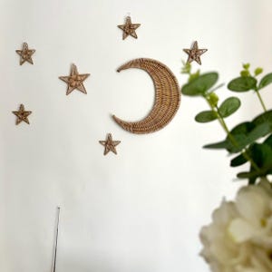 Op de afbeelding: Een set van gevlochten rotan wanddecoraties met een halve maan en sterren. De maan heeft een grote, gebogen vorm en de sterren zijn in verschillende maten. De decoratie heeft een natuurlijke bruine kleur en is bevestigd aan een witte muur. Ideaal voor een kinderkamer of slaapkamer.