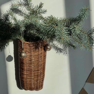 Könnte beinhalten: Ein brauner, geflochtener Weidenkorb zur Wandmontage, gefüllt mit grünen Tannenzweigen. Zwei kleine, runde, metallisch silberne Ornamente hängen an dünnen weißen Schnüren am Korb. Der Korb hat ein vertikales Flechtmuster und eine flache Rückseite.