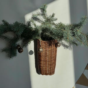 Pode incluir: Uma cesta de parede de vime castanho com um ramo de pinheiro verde e pequenos ornamentos prateados pendurados nela.