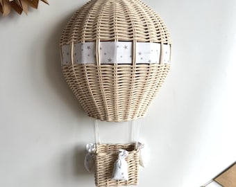 Op de afbeelding: Een wanddecoratie van een heteluchtballon van gevlochten wilg met een witte stoffen band met grijze sterren. De ballon heeft een mand van gevlochten wilg met twee kleine witte stoffen zakjes met grijze sterren.