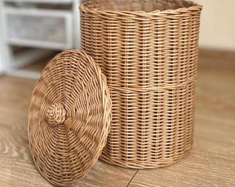 May include: A cylindrical wicker basket with a matching lid. The basket is made of woven light brown wicker and has a rounded lid with a small knob. The basket is designed for storage and is suitable for various rooms.