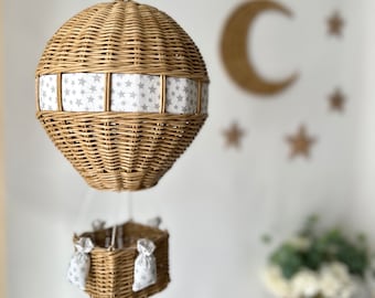 May include: A woven wicker hot air balloon mobile with a grey and white star-patterned band. The basket below has matching star-patterned fabric bags. The mobile is suspended, suggesting a nursery or children's room decor.