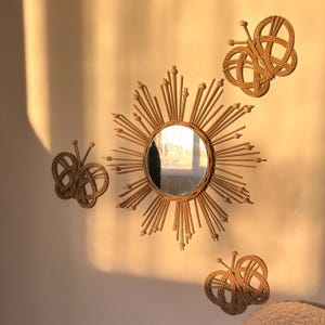 May include: Three wall decor pieces made of woven natural materials in the shape of a sunburst with a mirror in the center and two butterflies. The sunburst is brown and has a round mirror in the center. The butterflies are brown and have a natural wood finish.