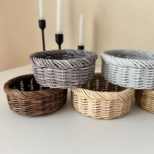 以下が含まれることがあります： 茶色、グレー、ベージュなど、さまざまな色の小さくて丸い織りバスケットのコレクション。 バスケットは白い表面に配置され、背景には黒いキャンドルホルダーと白いキャンドルがあります。 バスケットは天然素材で作られています。