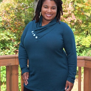 May include: Teal long-sleeved tunic with a cowl neck and three white buttons, worn with black leggings. The image is set outdoors with a wooden deck and trees in the background.