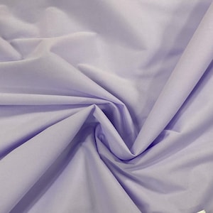 May include: A close-up shot of a light purple fabric with soft folds and subtle shadows. The fabric appears to be smooth and possibly cotton or a similar material, with a slight sheen. The overall color is a pale, muted lavender.