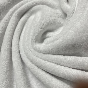 May include: Close-up of a white, soft, and fluffy fabric with a textured surface. The fabric is folded and draped, creating a swirling pattern.