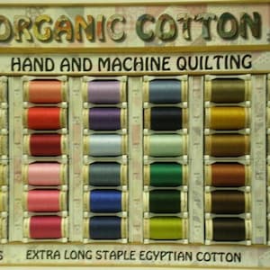 May include: A display of spools of organic cotton thread for hand and machine quilting. The spools are arranged in rows by color, with a variety of shades of white, beige, pink, red, purple, blue, green, brown, and gray. The spools are labeled "300 YDS" and "50 WT".