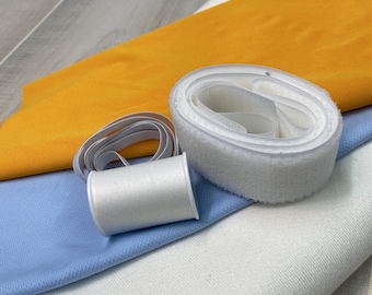 May include: A close-up of a white spool of thread, a white elastic band, and a white hook and loop fastener tape. The tape is rolled up and the elastic band is wrapped around the spool of thread. The items are on top of a blue fabric, a white fabric, and a yellow fabric.