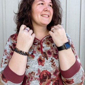 May include: A floral print hoodie with a burgundy zipper and cuffs. The sleeves are gray with a burgundy cuff. A black smartwatch is on the left wrist, and a beaded bracelet is on the right wrist. A purple ring is on the left hand.