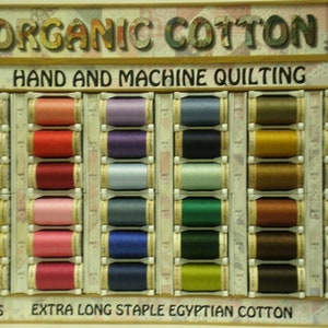 May include: A display of 50 weight organic cotton thread spools in various colors. The spools are arranged in rows and columns on a wooden shelf. The text "ORGANIC COTTON", "HAND AND MACHINE QUILTING", "300 YDS", "EXTRA LONG STAPLE EGYPTIAN COTTON", and "50 WT" is visible on the shelf.