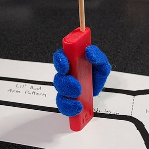 May include: A red rectangular object with a wooden stick inserted at the top is held by a blue felt hand. The object has the text "Lil' Bud" and "tj's-lab.com" printed on it. The background includes a white paper with text and black lines.