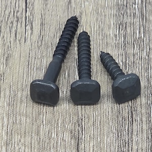 May include: Three black square-head wood screws of varying sizes are arranged on a light brown wooden surface. The screws have a dark, matte finish and a coarse thread. The heads are square with beveled edges.