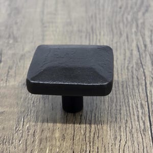 May include: Black square cabinet knob with a slightly raised, textured surface. The knob has a short, cylindrical post.