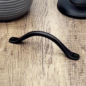 Puede incluir: Tirador de caj&oacute;n de metal negro con forma curva. El tirador tiene un dise&ntilde;o sencillo con bordes redondeados.