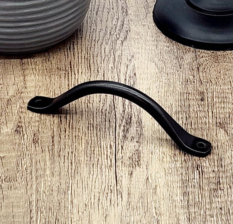 Puede incluir: Tirador de caj&oacute;n de metal negro con forma curva. El tirador tiene un dise&ntilde;o sencillo con bordes redondeados.