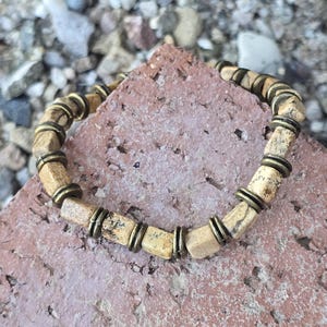 May include: A beaded bracelet featuring light tan, geometric-shaped beads with dark brown veining, separated by antique brass-colored metal rings. The bracelet is displayed on a textured, reddish-brown surface.