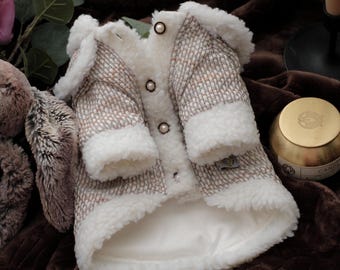 May include: A small dog coat with a tweed-like pattern in shades of brown and white, trimmed with fluffy white faux fur. The coat has a button-up front and a soft, white lining. The coat is designed to keep small dogs warm and stylish.