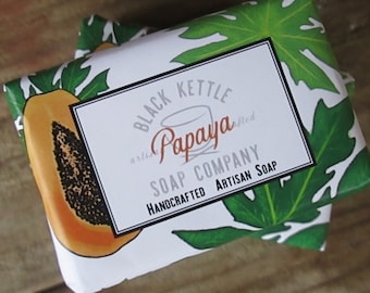 Può includere: Un sapone bianco con un'illustrazione di papaya e il testo "Black Kettle Artisan Papaya Soap Company Handmade Artisan Soap". Il sapone è avvolto in carta verde e bianca con un design a foglia di papaia.