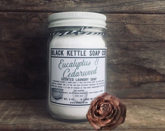 Op de afbeelding: Een glazen pot met een wit deksel gevuld met witte wasmiddel. Het etiket op de pot luidt "BLACK KETTLE SOAP CO Eucalyptus & Cedarwood SCENTED LAUNDRY SOAP 18 LOADS www.blackkettlesoap.co".