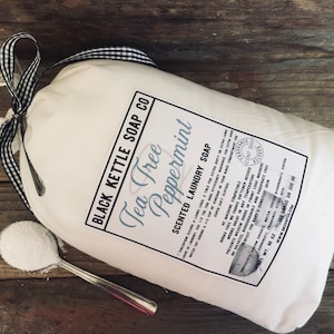 May include: A white canvas bag with a black and white checkered ribbon tied around it. The bag contains a white laundry soap powder. The label on the bag reads "Black Kettle Soap Co. Tea Tree Peppermint Scented Laundry Soap". The label also includes a list of ingredients and instructions for use. The bag is sitting on a wooden surface.
