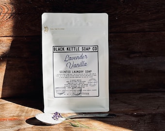Op de afbeelding: Een witte zak met een zwart label waarop staat "Black Kettle Soap Co. Lavender Vanilla Scented Laundry Soap". De zak ligt op een houten oppervlak met een lepel met witte wasmiddel en lavendelbloemen.