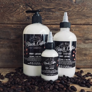 May include: Three bottles of Black Kettle Black Coffee Soap Company body lotion. The bottles are white with black labels and feature a coffee cup logo. The bottles are sitting on a wooden surface surrounded by coffee beans.