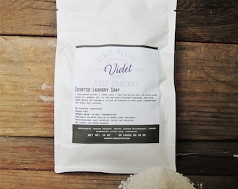 Op de afbeelding: Een witte hersluitbare zak met een zwart label waarop staat "Black Kettle Violet Scented Laundry Soap Company". Het label bevat ook de ingrediënten en het nettogewicht van de zeep: 14 oz, 28 ladingen of 56 HE. De zak ligt op een houten oppervlak met een stapel witte waspoeder ervoor.
