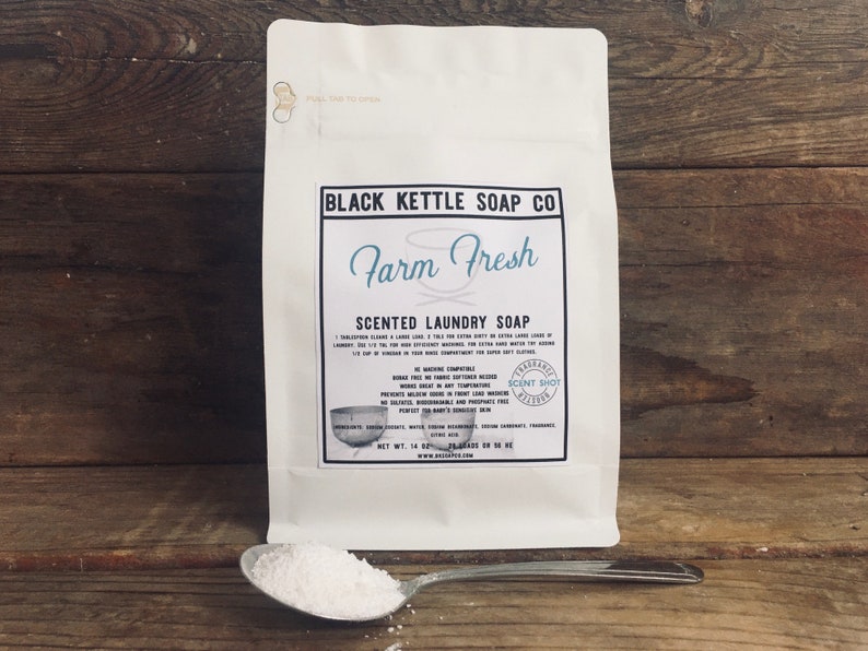 Op de afbeelding: Een witte zak met Farm Fresh geparfumeerde wasmiddel van Black Kettle Soap Co. De zak ligt op een houten oppervlak met een witte lepel gevuld met waspoeder.