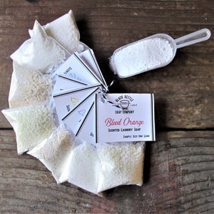 May include: A set of six small white bags filled with white laundry soap granules. Each bag has a small tag with the scent name. The tag for the bag in the centre reads "Blood Orange Scented Laundry Soap". The text "Black Kettle Soap Company" is on the tag. A white scoop with white laundry soap granules is in the top right corner of the image.