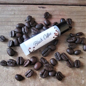 May include: A white tube of lip balm with a black cap and a label that says "Black Coffee". The tube is surrounded by coffee beans on a wooden surface.