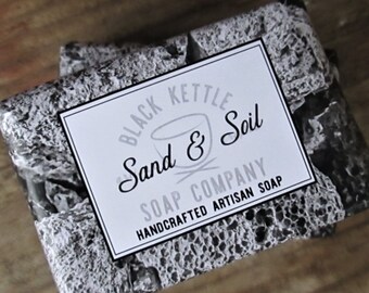 Puede incluir: Una pastilla de jabón con un diseño estampado en blanco y negro. La etiqueta dice "Black Kettle Sand & Soil Soap Company Handcrafted Artisan Soap".
