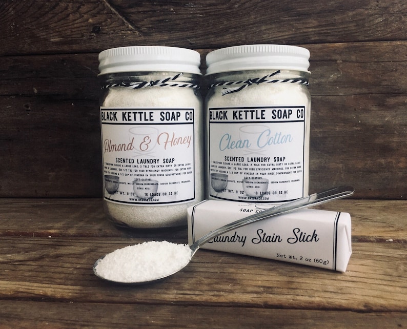 May include: Two glass jars filled with white laundry soap, labeled "Black Kettle Soap Co." with the scents "Almond & Honey" and "Clean Cotton". A silver spoon holds a scoop of the soap and a white bar labeled "Laundry Stain Stick" is in the foreground.