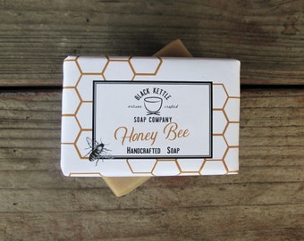 May include: A bar of soap in a white box with a gold honeycomb pattern. The box has a black label with the text "Black Kettle Soap Company" and "Honey Bee Handcrafted Soap".