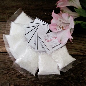 May include: Eight small white plastic bags filled with white laundry soap beads. The bags are arranged in a circle on a wooden surface. A white label with black text reads "Black Kettle Flower Box Soap Company Scented Laundry Soap". A pink flower is in the background.