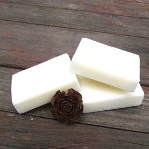 Peut inclure: Trois barres de savon blanc empil&eacute;es les unes sur les autres sur une surface en bois. Une petite pomme de pin brune se trouve devant le savon.
