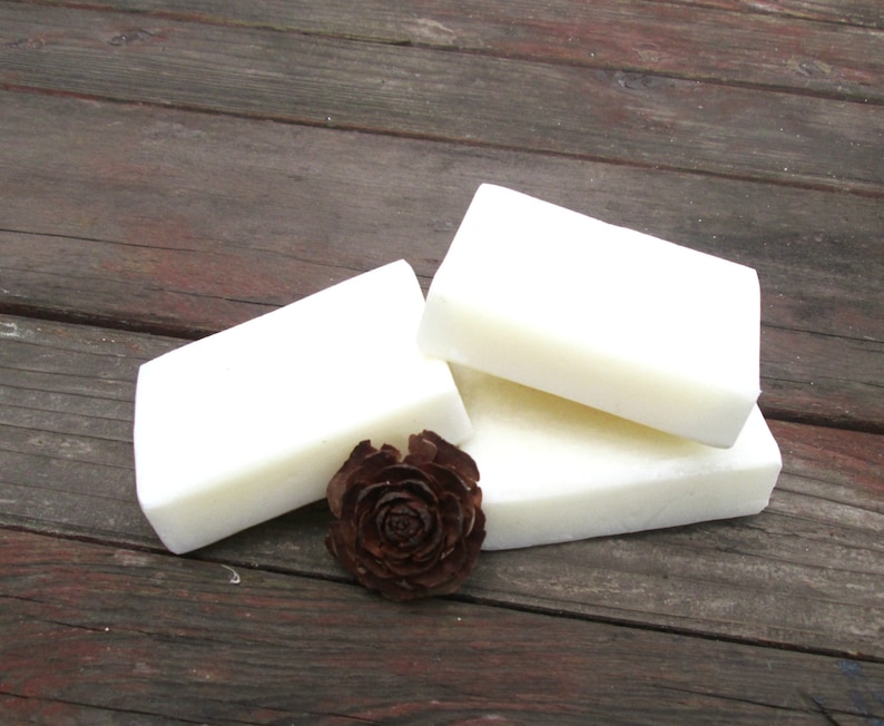 Peut inclure: Trois barres de savon blanc empil&eacute;es les unes sur les autres sur une surface en bois. Une petite pomme de pin brune se trouve devant le savon.