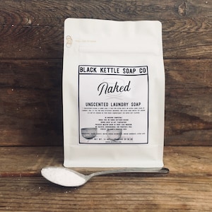 Può includere: Un sacchetto bianco di sapone per bucato non profumato con il nome del marchio "Black Kettle Soap Co." e il nome del prodotto "Naked" stampato sul davanti. Il sacchetto è su una superficie di legno. Un piccolo cucchiaio con polvere bianca è in primo piano.