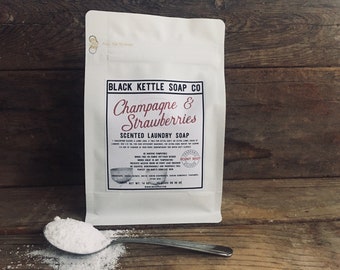 Op de afbeelding: Een witte zak "Champagne & Strawberries" geurende wasmiddel van Black Kettle Soap Co. Een metalen lepel bevat een kleine hoop van het witte poeder. De zak staat op een houten oppervlak.