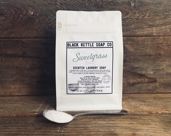 May include: A white pouch of Black Kettle Soap Co. Sweetgrass scented laundry soap with a label detailing usage instructions and ingredients. A metal spoon filled with white powdered laundry soap rests in front of the pouch. Net weight 14 oz.