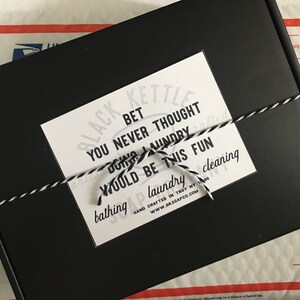 May include: A black box with a white label that reads "BLACK KETTLE BET YOU NEVER THOUGHT DOING LAUNDRY WOULD BE THIS FUN bathing laundry cleaning HAND CRAFTED IN TROY NY 12180 www.BKSOAPCO.COM". The box is tied with a black and white string.