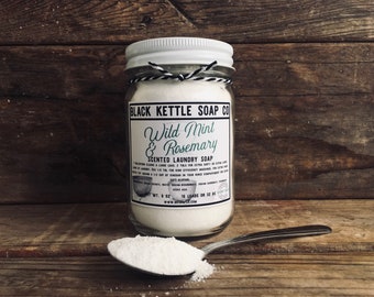 May include: A glass jar filled with white laundry soap powder. The jar has a white lid and a label that reads "Black Kettle Soap Co. Wild Mint & Rosemary Scented Laundry Soap. WT. 8 oz. 18 Loads or 32 oz. www.blackkettlesoapco.com".