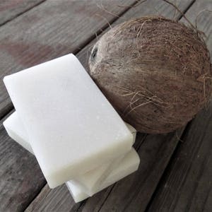 Peut inclure: Une pile de trois barres de savon rectangulaires blanches &agrave; c&ocirc;t&eacute; d'une noix de coco brune sur une surface en bois.