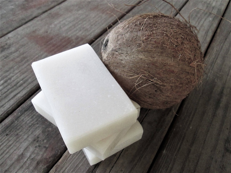 Peut inclure: Une pile de trois barres de savon rectangulaires blanches &agrave; c&ocirc;t&eacute; d'une noix de coco brune sur une surface en bois.
