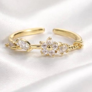 May include: A gold-colored adjustable ring with a floral design. The ring features a central flower with clear, round-cut stones and smaller, leaf-shaped stones on either side. The ring is set against a white, silky background.