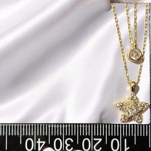 May include: A gold double-layered necklace featuring a small round solitaire pendant and a larger star-shaped pendant encrusted with clear stones. A black ruler is visible, showing the star pendant measures approximately 0.5 inches in width.