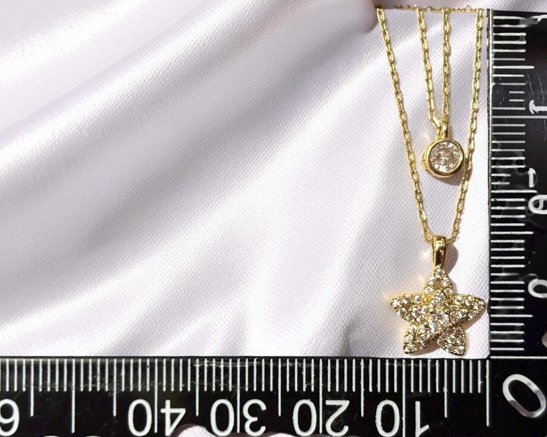 May include: A gold double-layered necklace featuring a small round solitaire pendant and a larger star-shaped pendant encrusted with clear stones. A black ruler is visible, showing the star pendant measures approximately 0.5 inches in width.