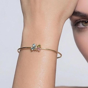 May include: A gold-toned bracelet with a butterfly design. The butterfly has blue and pink gemstones. The bracelet is worn on a wrist, with a person's face visible in the background.