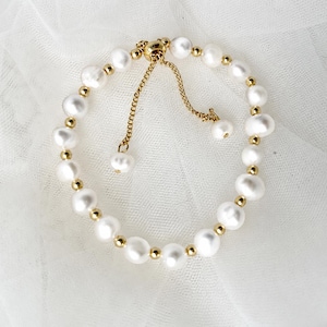 May include: A bracelet featuring white freshwater pearls alternating with small gold-toned metal beads. The piece includes a gold-toned adjustable sliding chain mechanism with two dangling pearl-tipped ends to secure the fit around the wrist.